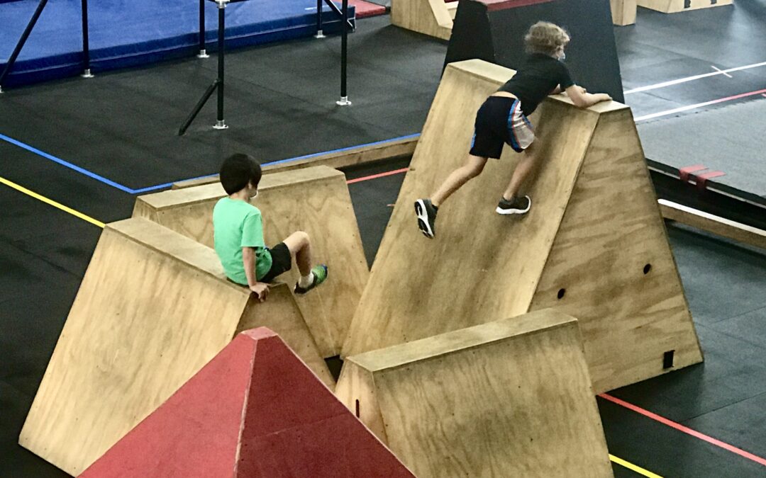 Two children in athletic wear navigate a wooden climbing structure in an indoor activity center, reminiscent of a parkour competition.