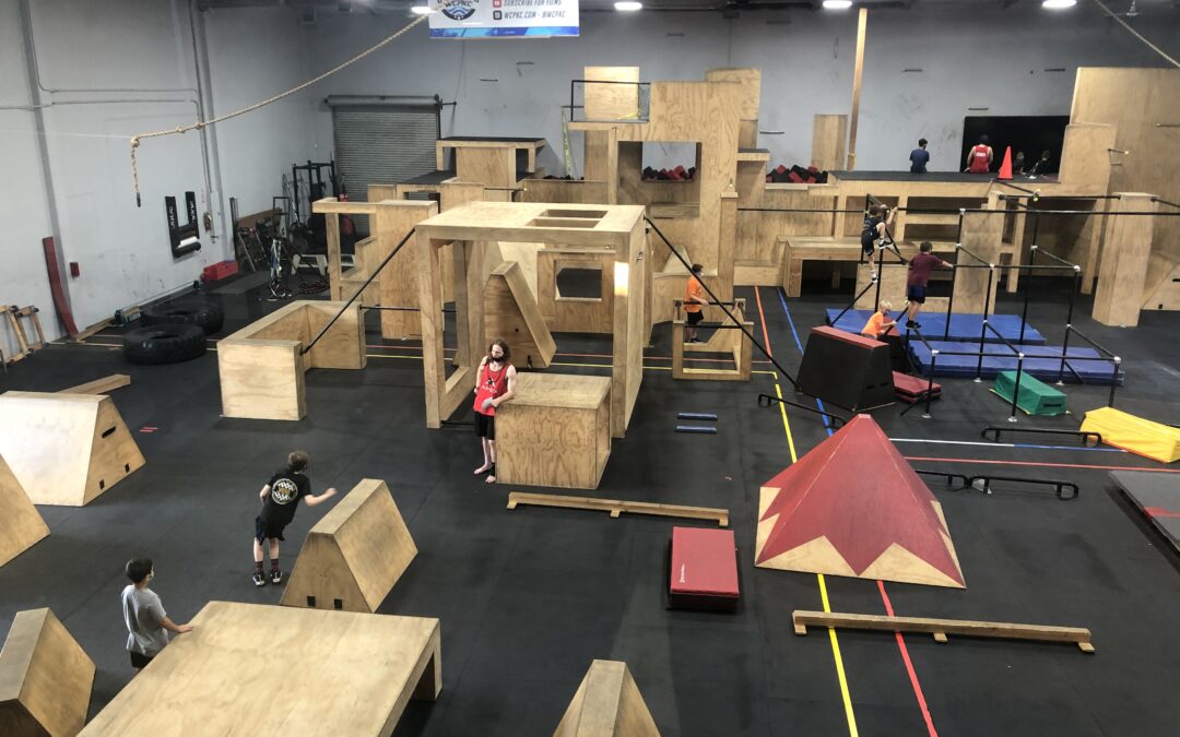 A large indoor parkour gym in California with various wooden obstacles. Several people, both children and adults, are practicing parkour activities on the structures. The environment is well-lit and spacious, offering classes for all ages and skill levels.